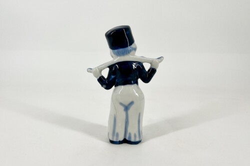 Ceramic figurine seen from behind, wearing a dark blue coat and tall hat with white scarf across the shoulders.