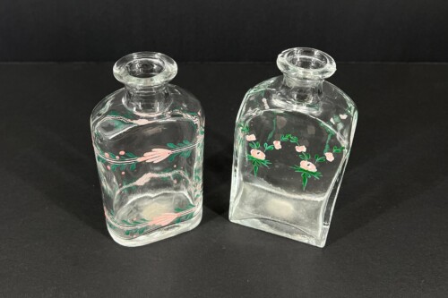 Two clear glass bottles with delicate pink floral patterns and green leaves, set on a black surface against a dark backdrop.