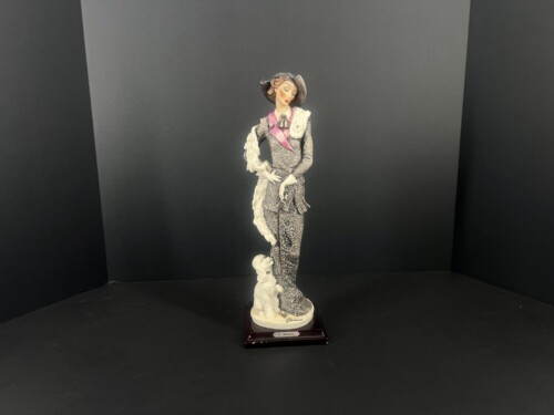 Porcelain figurine of a 1920s-era woman in a gray patterned dress with pink scarf, black hat, and white gloves, standing on a pedestal with a decorative white shawl draped over one arm against a dark backdrop.