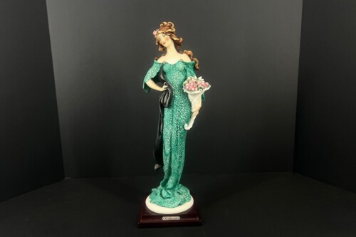 Statue of a woman in a sparkling green off-shoulder gown holding a bouquet, with a black shawl draped over one arm, standing on a white base with a wooden plaque.