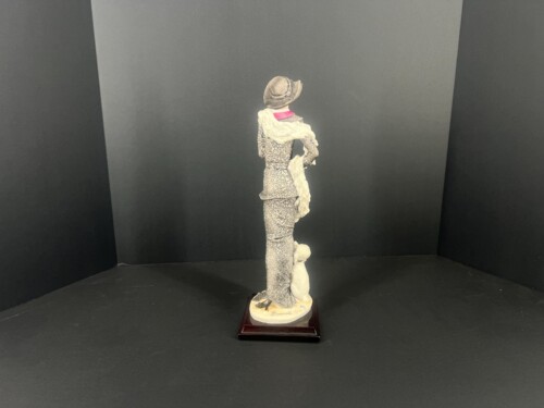Back-view statue: a small female figure in a textured patterned dress, hat, pink collar, and cream scarf, standing on a wooden pedestal in a black display corner.