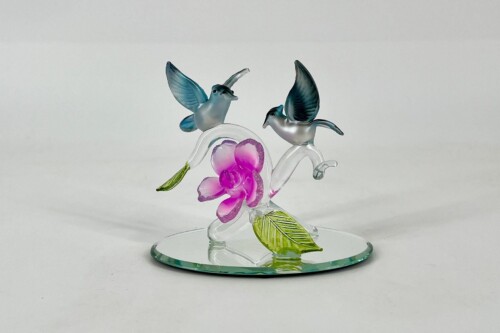 Glass sculpture of two blue hummingbirds hovering above a pink orchid on a clear curved stem, mounted on a round glass base.