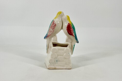 Ceramic sculpture of two colorful parrots perched together, forming a heart-like shape, with yellow heads and multicolored wings on a white pedestal