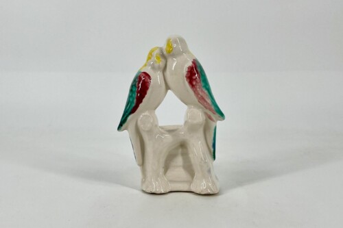 Ceramic figurine of two parrots perched together, facing each other with beaks touching; white base, teal and red wings, yellow heads.