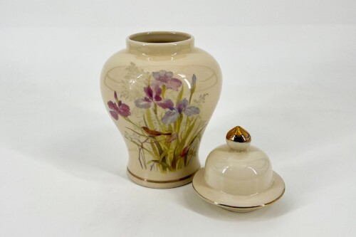 Cream-colored ceramic vase with purple and yellow flowers, placed beside its matching lid with a gold knob.