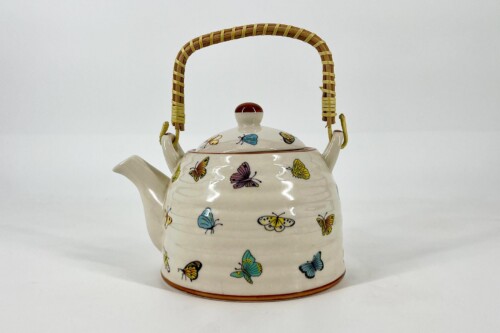 White ceramic teapot covered in colorful butterfly illustrations with a woven wicker handle and red knob on the lid, on a plain background