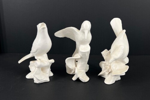 Three white ceramic bird sculptures displayed on a dark surface, with left bird upright, center bird with wings outstretched, and right bird standing with a raised tail detail on a sculpted base.