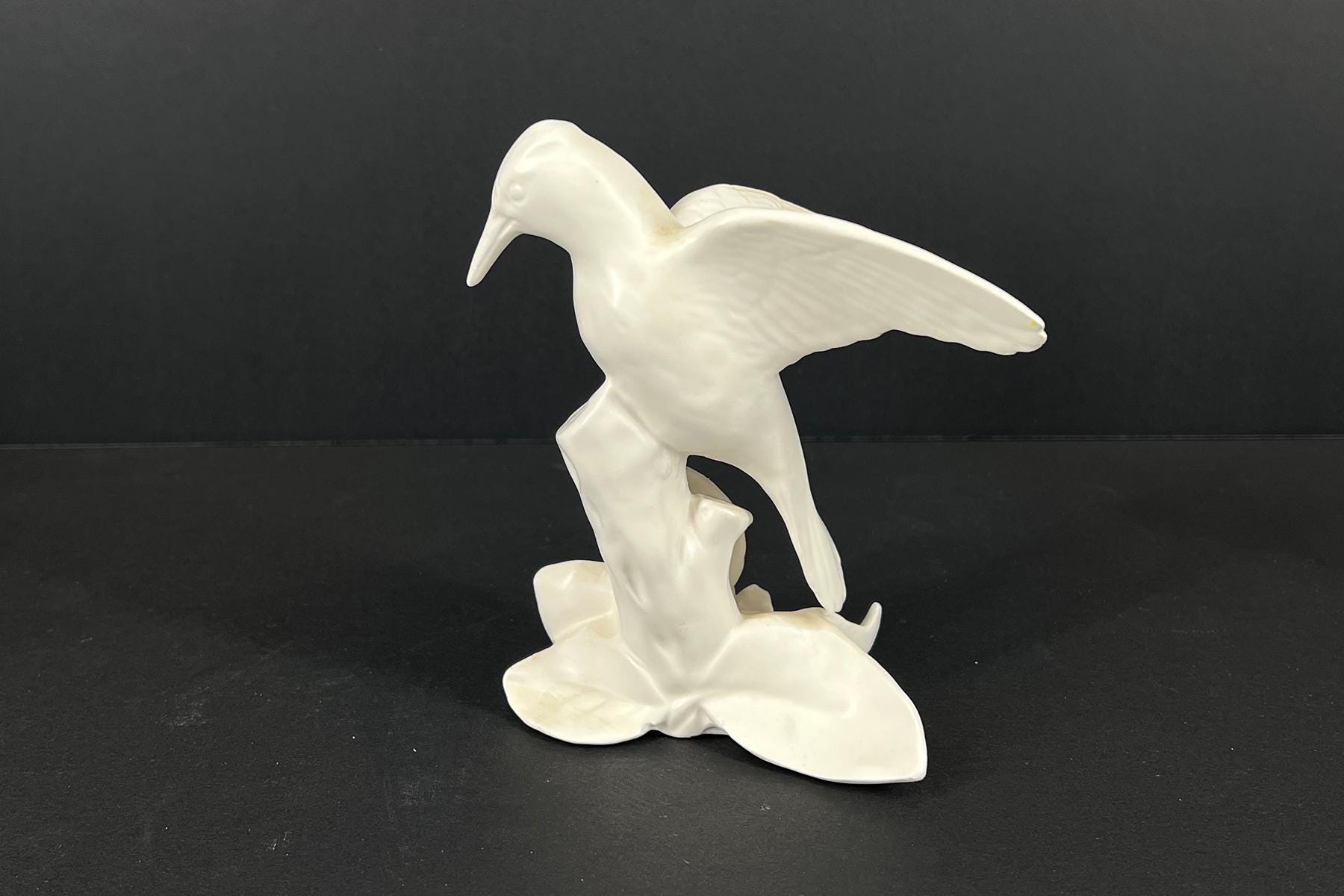 White ceramic bird sculpture perched on a rocky base, with wings tucked to its sides and a long beak visible