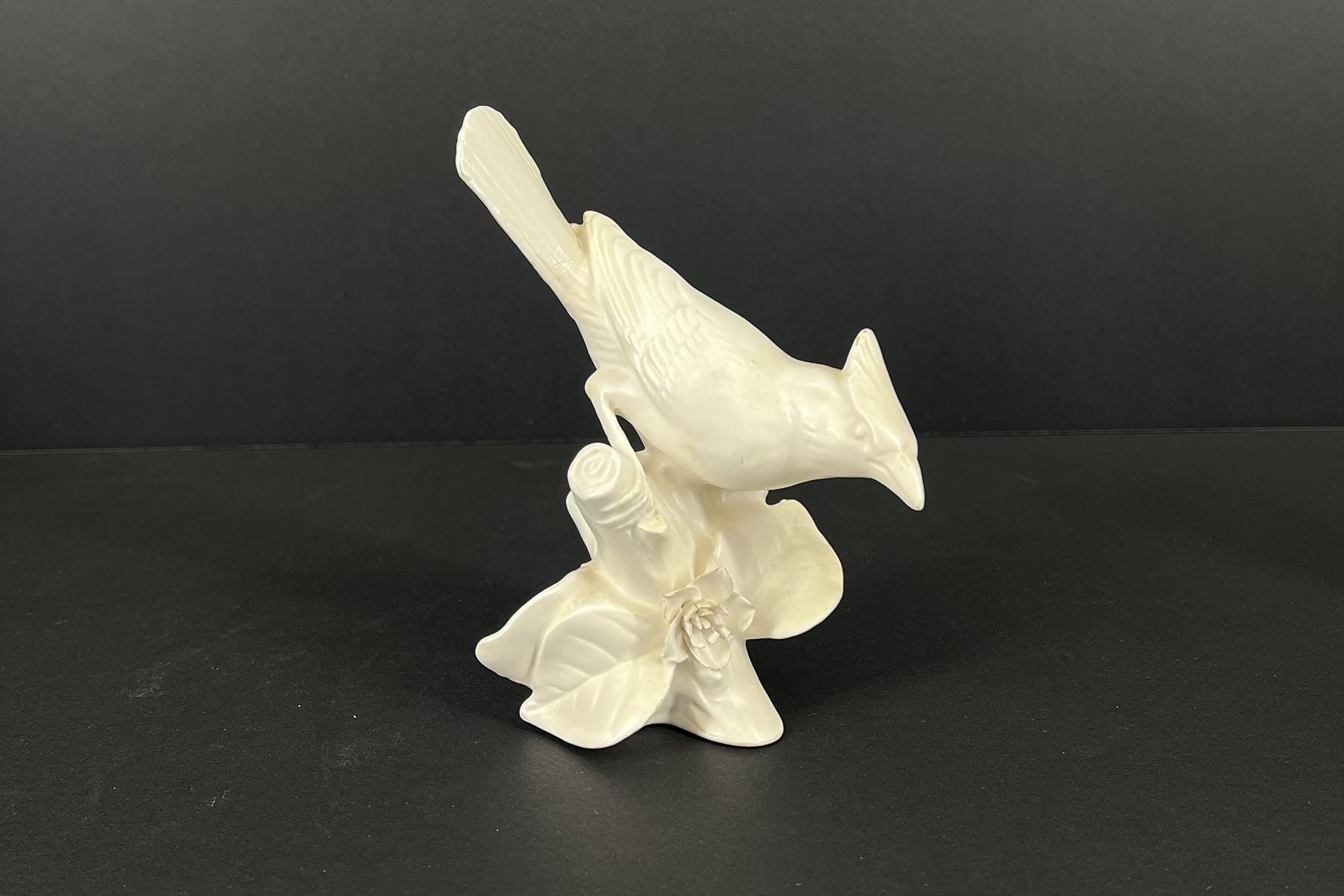 White ceramic sculpture of a bird with a long beak perched on a floral pedestal, against a dark background