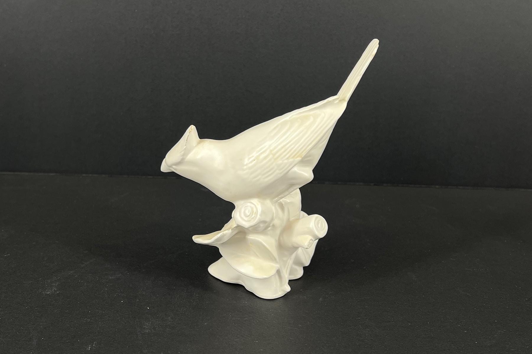 White ceramic bird figurine with a long tail perched on a decorative, spiral-based base against a dark background.