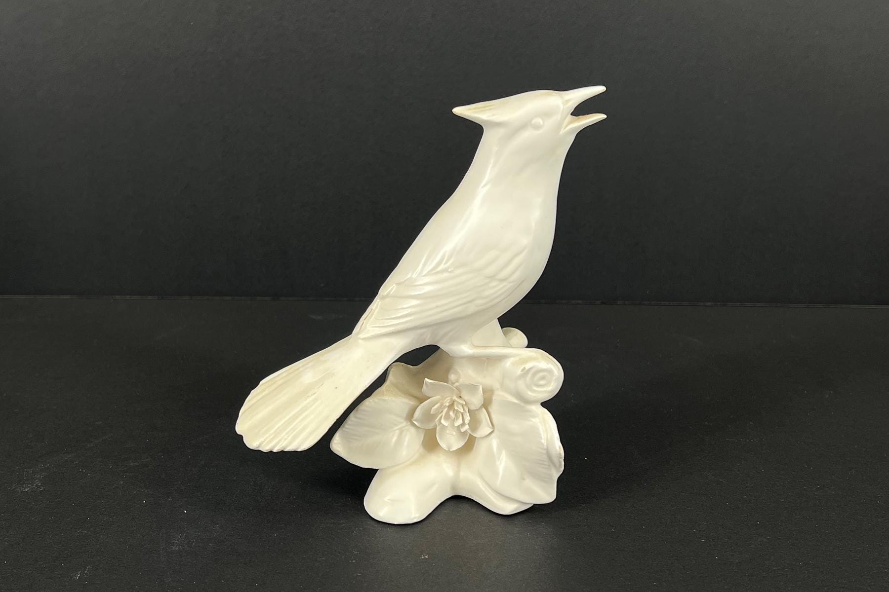 White ceramic bird statue perched on a floral pedestal against a black background.