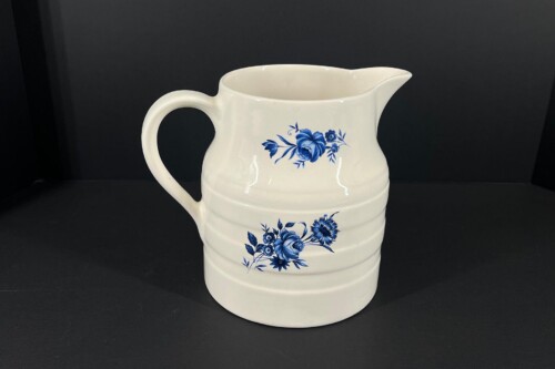 White ceramic pitcher with a blue floral design and a curved handle against a dark background, ready to pour.