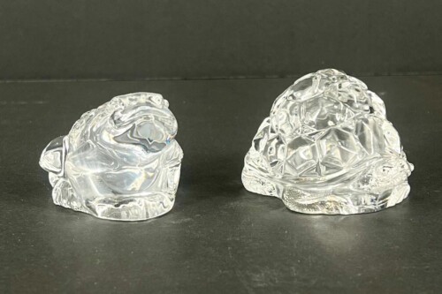 Two clear glass sculptures with jagged, faceted surfaces sit on a dark surface against a black background.