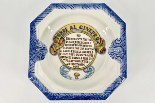 Octagonal ceramic plate with a blue patterned rim and a central circular plaque containing Italian text in yellow.