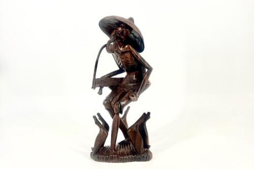 Bronze sculpture of a slender figure wearing a wide-brimmed hat, perched on a base with outstretched hands, appearing to blow into a curved horn-like object