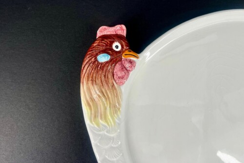 Ceramic plate with a rooster-shaped handle on the left edge, showing a brown and red rooster head with an orange beak against a dark background.
