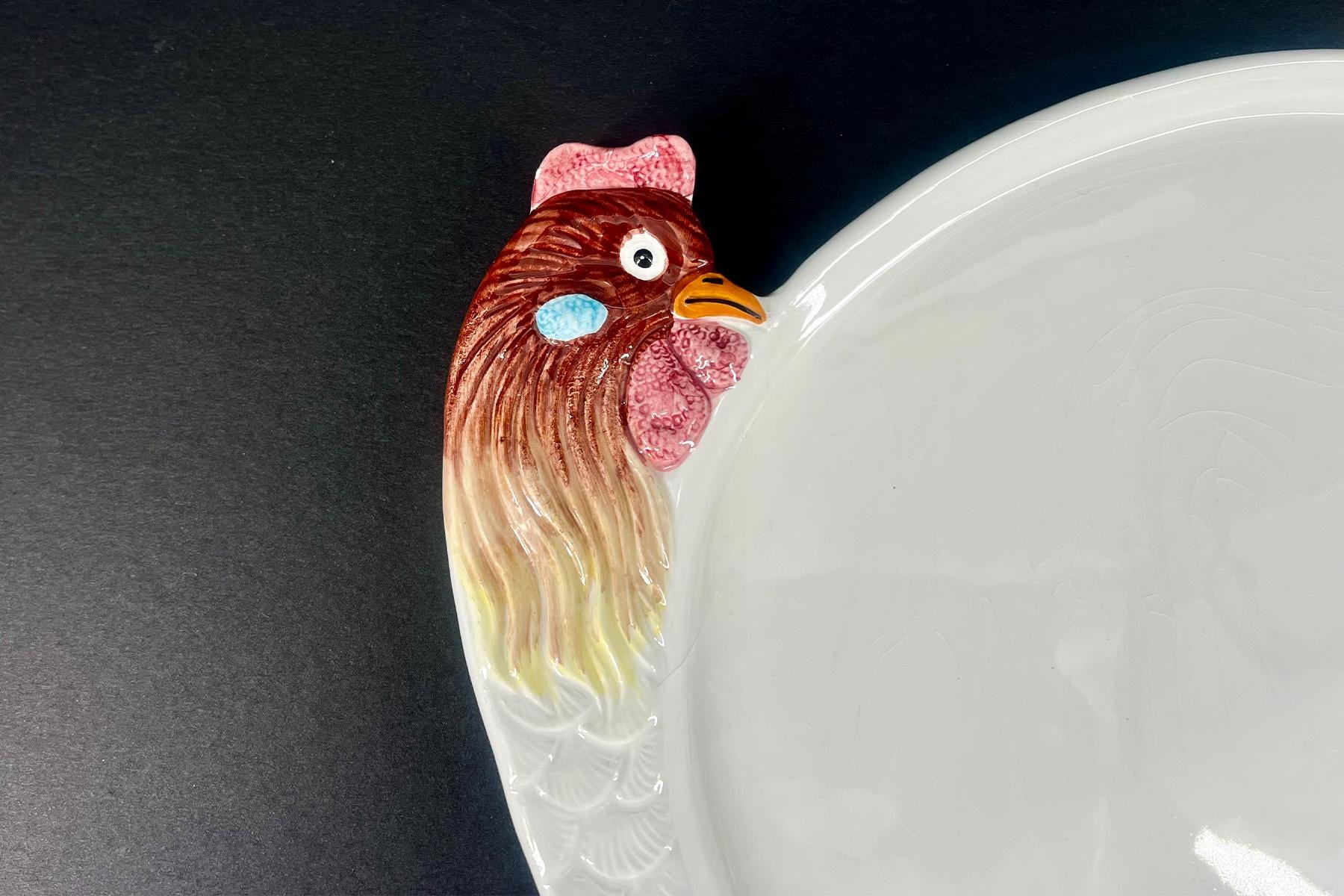 Ceramic plate with a rooster-shaped handle on the left edge, showing a brown and red rooster head with an orange beak against a dark background.