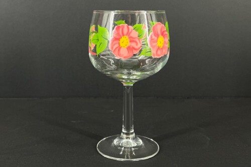 Clear stemmed glass decorated with bright pink flowers and green leaves against a dark background.