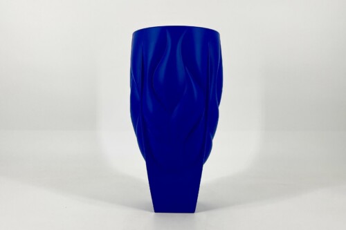 Tall cobalt-blue vase with flowing, wave-like ridges standing on a white surface against a plain white background, photography-focused still life.