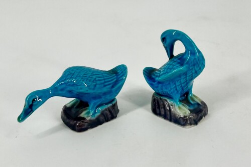 Two blue ceramic bird figurines on dark rocky bases, one bending its neck toward the ground and the other standing upright nearby.