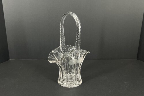 Clear cut-crystal basket vase with a tall loop handle and etched floral patterns set against a dark background.