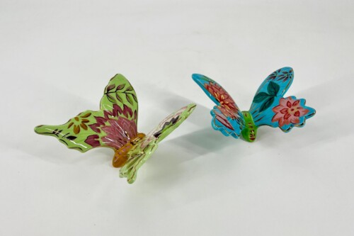 Two colorful ceramic butterfly figurines on a white surface: one green with pink markings, the other blue with floral patterns, appearing to fly apart.