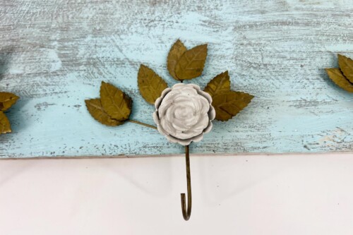 Decorative metal hook with a white ceramic rose centerpiece on a blue distressed wooden backdrop, surrounded by dried autumn leaves