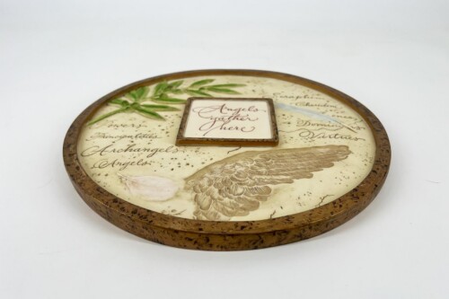 Round decorative plaque with parchment-like background, a green sprig, and a central tile that reads 'Angels gather here' amid a feathered wing motif.