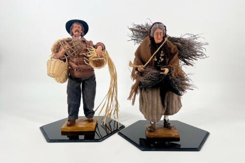 Two handmade wooden dolls in rustic outfits stand on black bases; a man with a hat carries woven baskets and straw a lot of straw pieces in his arms?