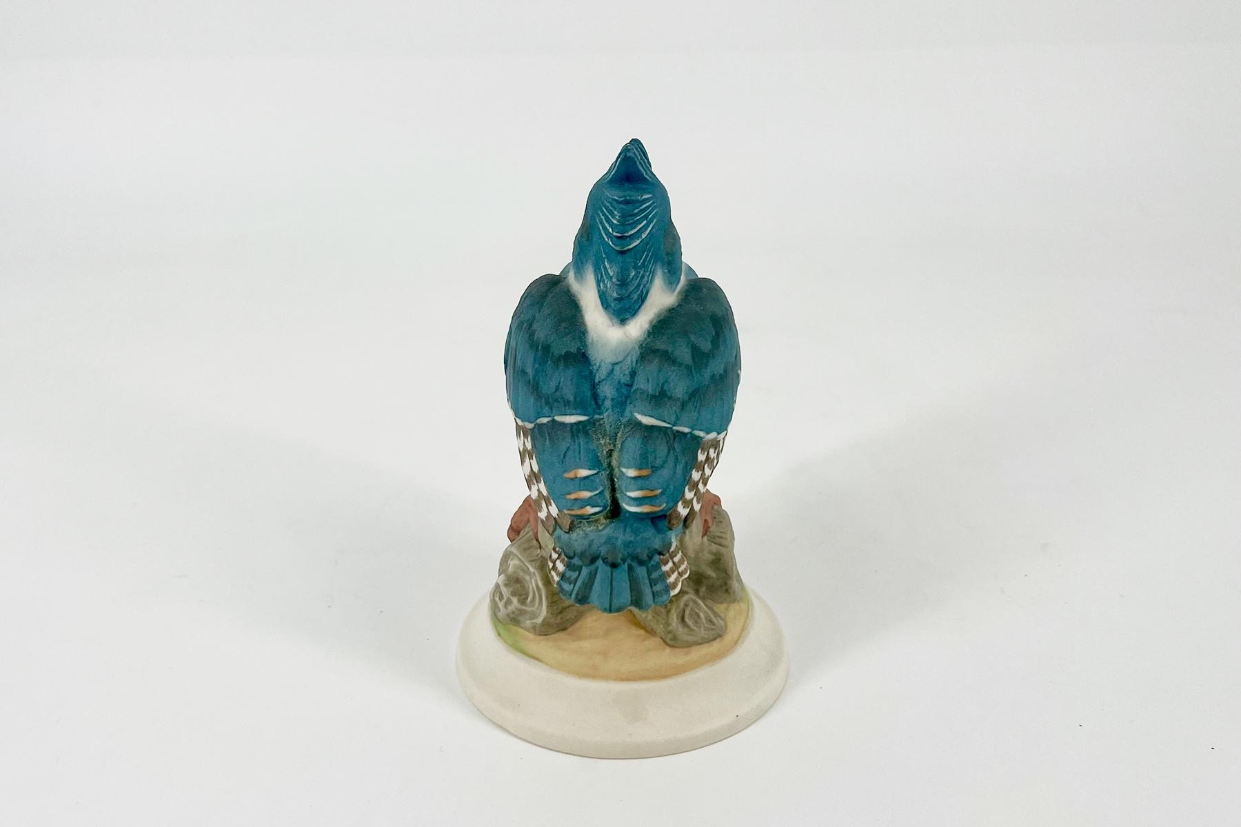 Blue parrot figurine viewed from behind, perched on a round beige pedestal.
