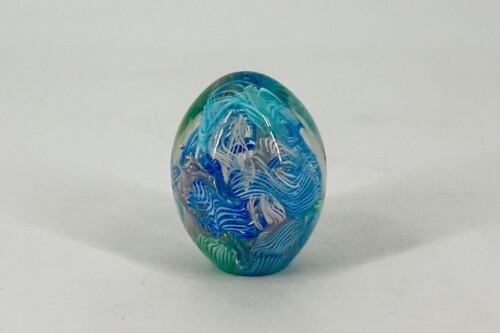 Glass art egg with blue and green swirls, standing on a white surface against a plain light background