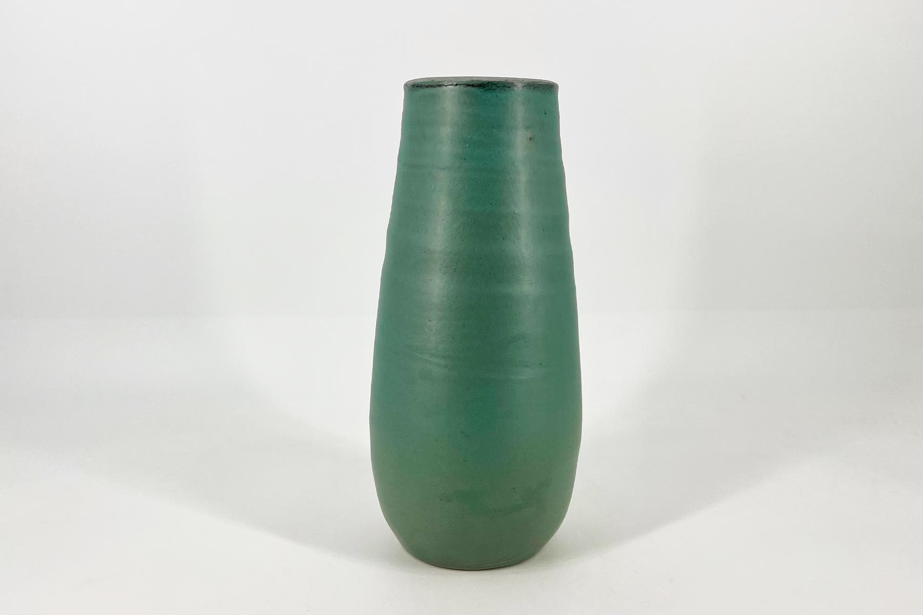 Tall green ceramic vase with a tapered cylindrical body on a white surface in a pale studio backdrop.