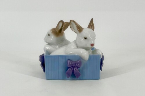 Two ceramic white rabbits in a blue gift box with a purple ribbon, positioned cuddled together as a decorative figurine set