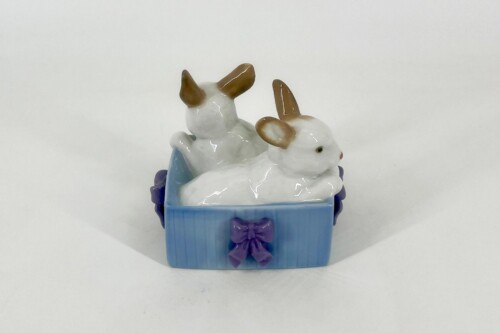 Ceramic figurine of two white rabbits with brown ears in a blue basket tied with a purple bowy accessory