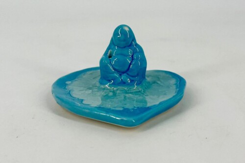 Blue clay sculpture of a seated, abstract figure on a matching blue round base, posed with an arm raised.