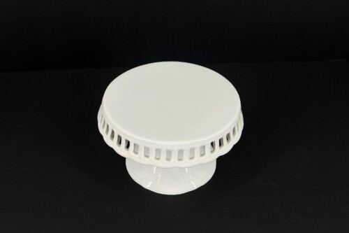 White ceramic cake stand with a smooth round top and a decorative perforated edge, set against a black background.