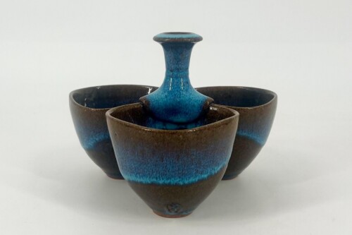 Three brown ceramic bowls with blue glaze form a nested, stem-like centerpiece with a turquoise-blue vase rising from the middle.