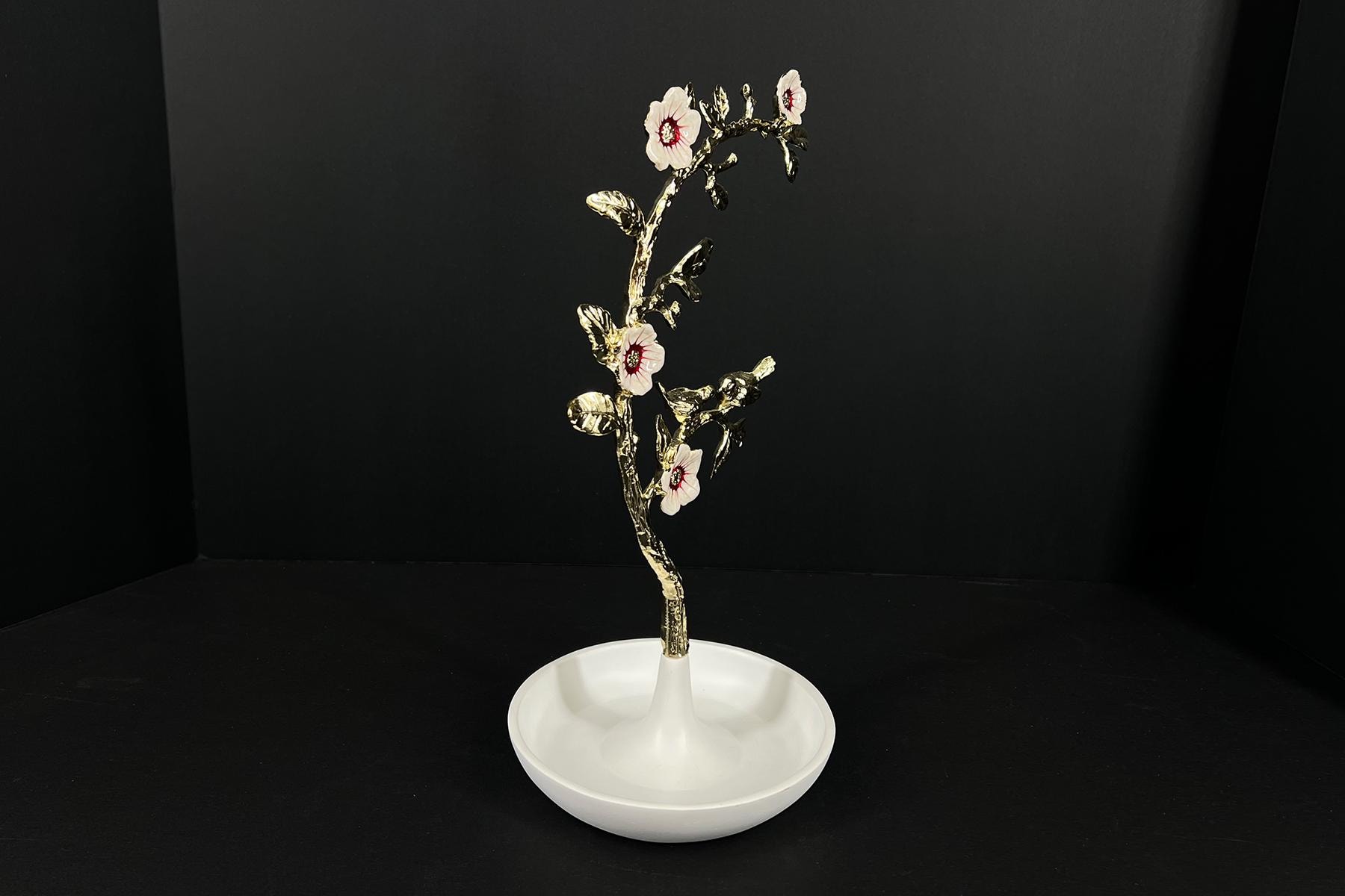 Gold-toned decorative flowering branch with pale pink centers, mounted on a white pedestal against a black background.