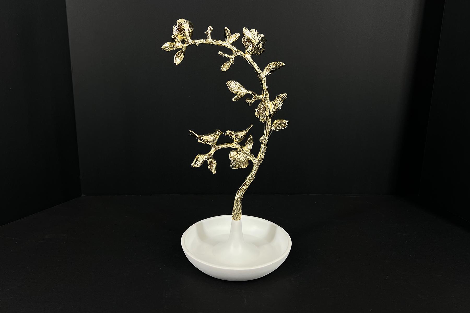 Gold-painted floral sculpture on a white pedestal vase against a black backdrop, curved branch with blossoms.