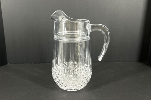 Clear cut-crystal pitcher with a curved handle, lid, and diamond-cut base against a dark background.