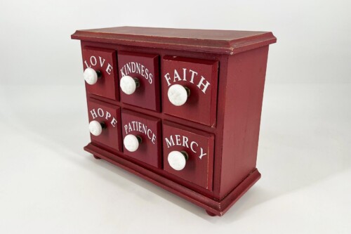 Red vintage wooden cabinet with six small drawers, each labeled Love, Kindness, Faith, Hope, Patience, Mercy, and white knobs on the fronts.