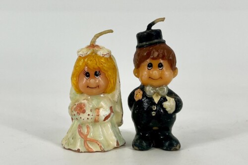 Two small wedding cake toppers: a smiling bride in a white dress with blond hair and a groom in a black suit with a bow tie, against a plain background.