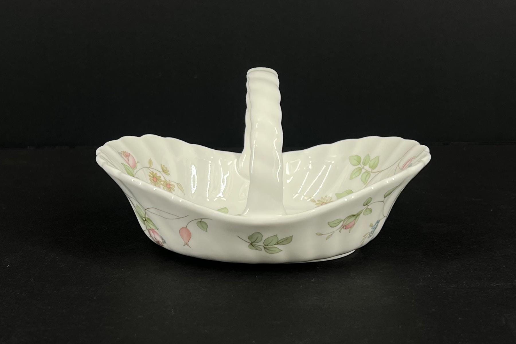 White porcelain basket with a tall, ridged handle and scalloped floral rim, decorated with pastel vines and pink flowers (decorative serving piece).