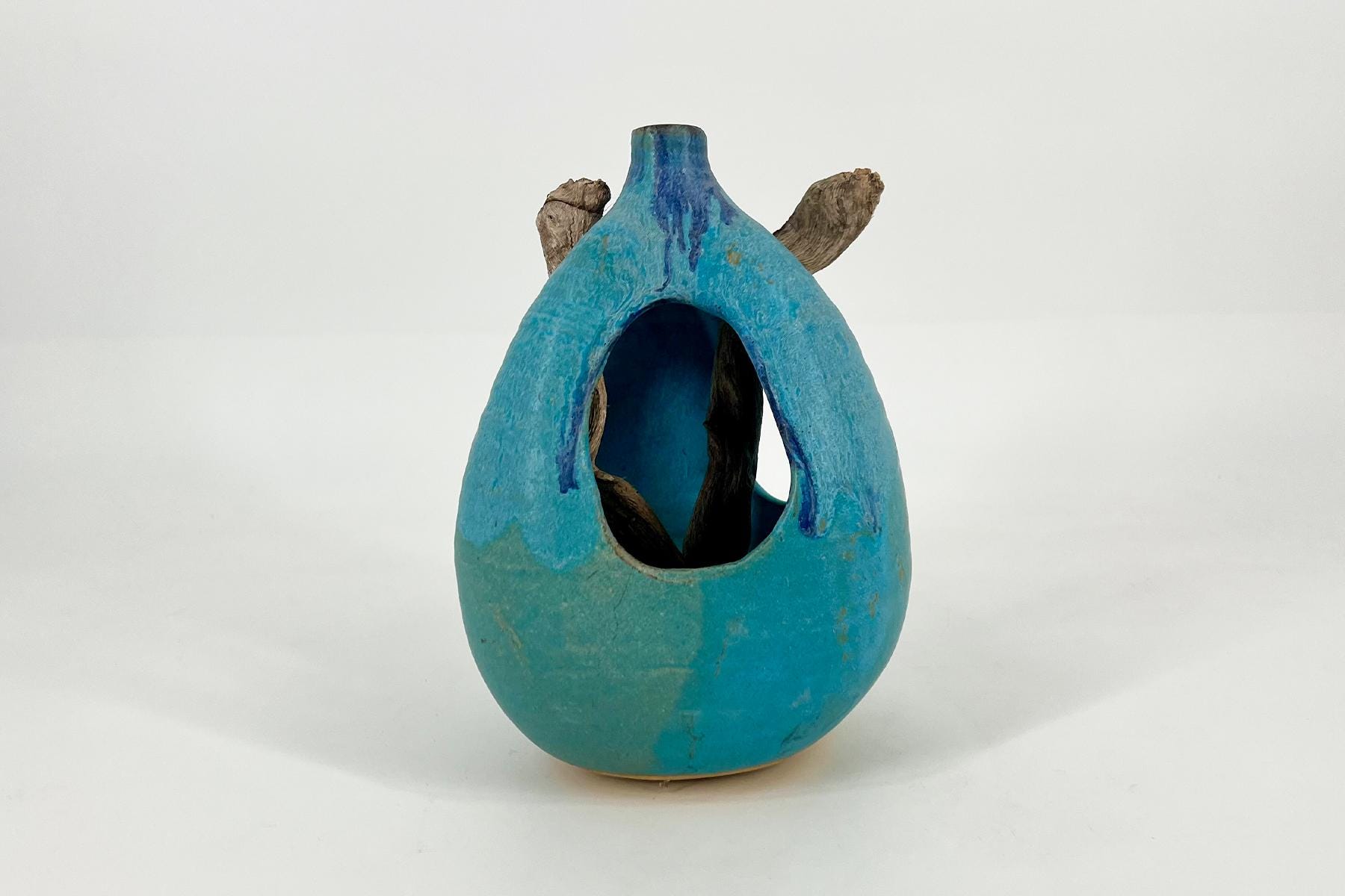 Blue ceramic hollow sculpture shaped like a teardrop birdhouse with a large front opening and brown twig-like protrusions.