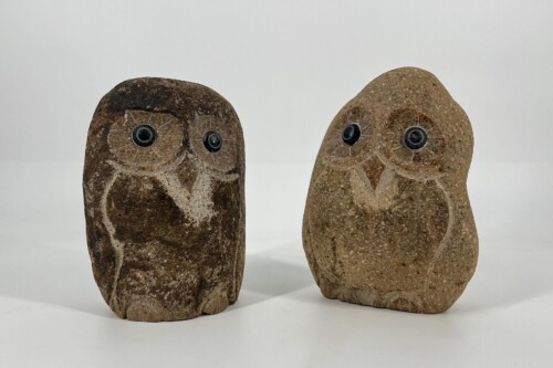 Two carved stone owl figurines standing side by side on a white surface, each with round black eyes and etched feather details.