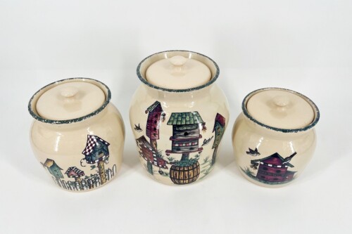 Three cream-colored lidded ceramic jars with hand-painted village scenes and teal-rimmed edges, arranged in a row on a white surface